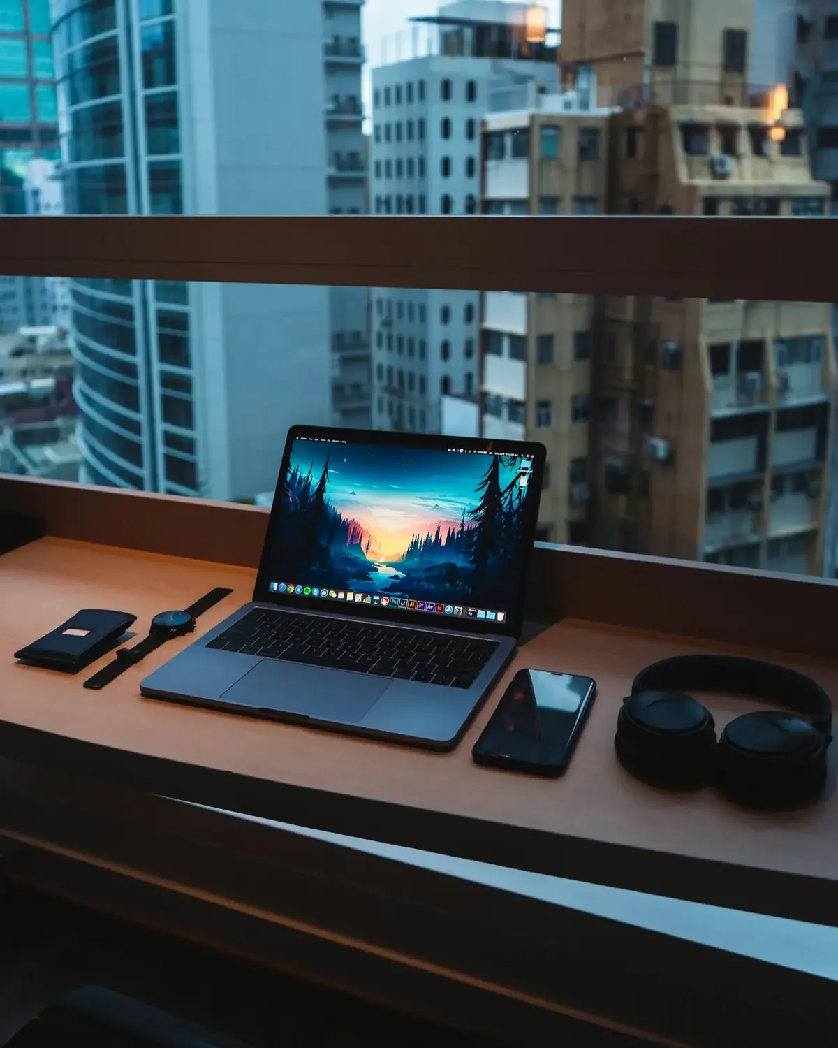 MacBook Pro open on a wooden desk by a city window, next to a phone, watch and headphones