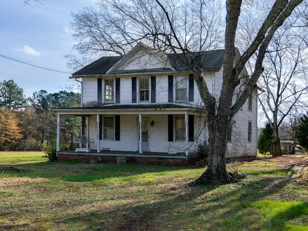 Older white two-storey farmhouse with a wraparound porch and peeling paint, the kind of home often sold as-is